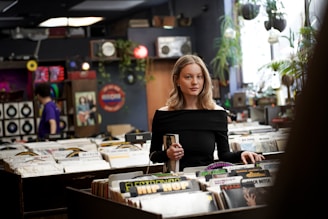 A young woman stands in a record store, browsing through vinyl records. She is holding a record in her hand and wearing an off-shoulder black top. The store is filled with rows of vinyls, and the walls are decorated with music-themed decor and plants.