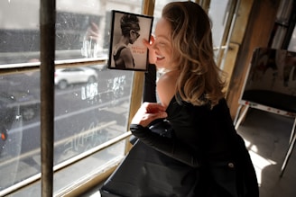 A person wearing a black outfit is leaning against a window with sunlight streaming in, while smiling and looking at a framed image on the wall. The image features a black and white photo of a woman with sunglasses and an updo hairstyle, with the text 'Fashion in Film.' The bright environment and casual pose create a relaxed and joyful atmosphere.