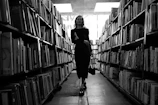 A cinematic black and white photo of a solitary figure walking through an ancient library aisle.