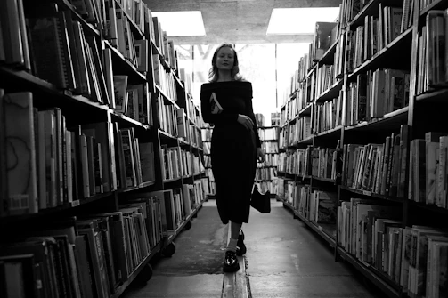 A cinematic black and white photo of a solitary figure walking through an ancient library aisle.