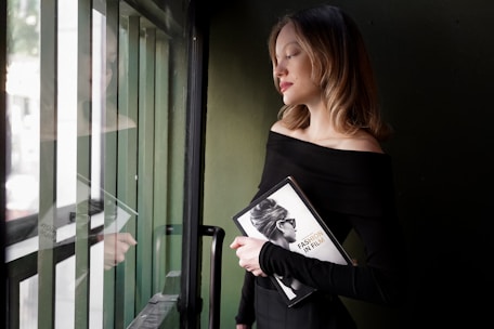 A woman stands near a window, holding a book titled 'Fashion in Film' with an image of a lady wearing sunglasses on its cover. She has light brown hair and is dressed in a sleek, off-the-shoulder black outfit. Her expression is calm and serene, and she is gazing to the side, capturing a reflective and relaxed atmosphere.