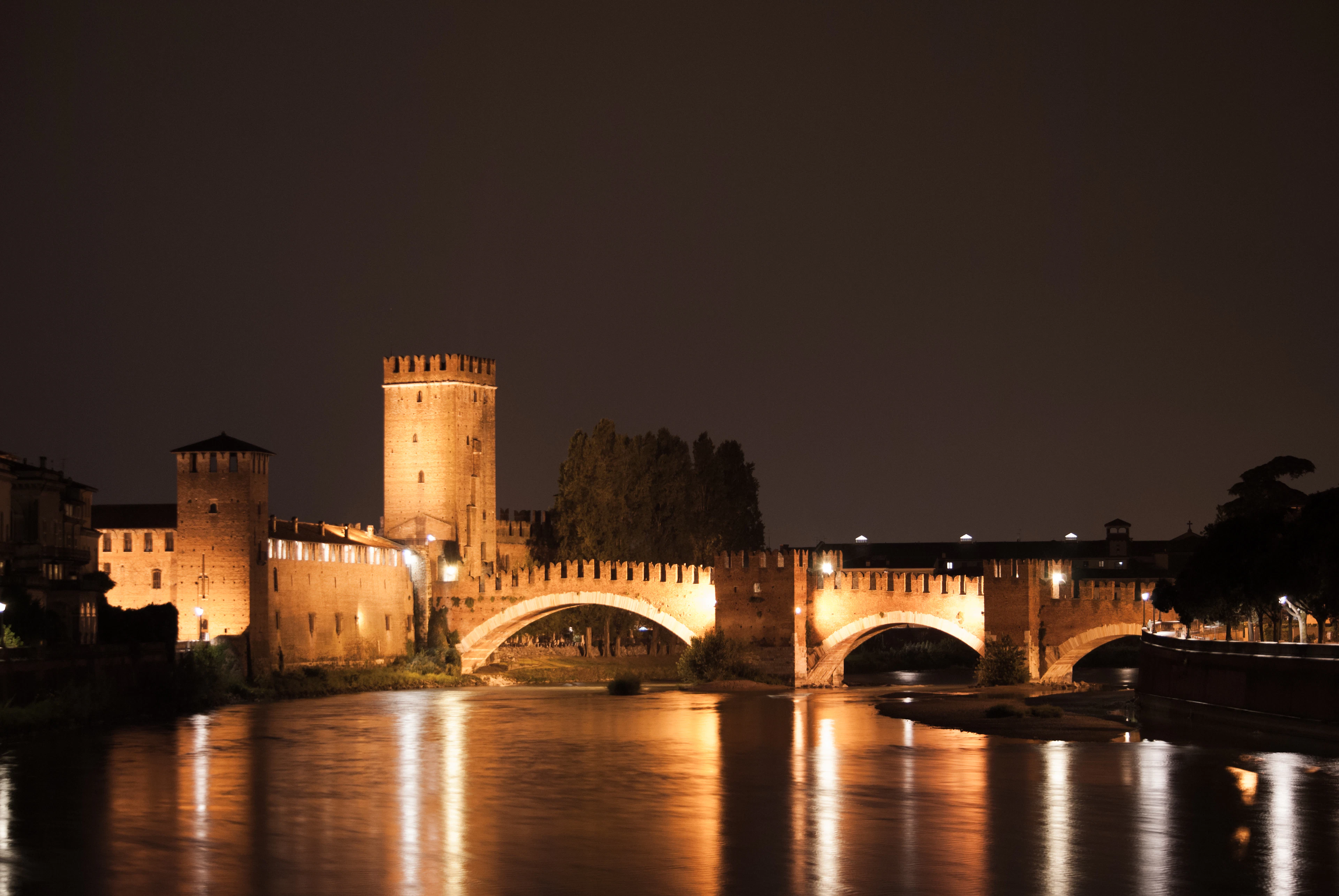 The brick keep and battlemented arches of Castelvecchio and its Ponte Scaligero in Verona lit warm gold at night, their reflection stretching in long ribbons across the dark Adige.