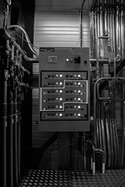 A grayscale image of an industrial control panel with numerous buttons and switches mounted on a wall surrounded by various pipes and metal conduits.