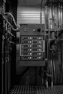 A grayscale image of an industrial control panel with numerous buttons and switches mounted on a wall surrounded by various pipes and metal conduits.