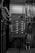 A grayscale image of an industrial control panel with numerous buttons and switches mounted on a wall surrounded by various pipes and metal conduits.
