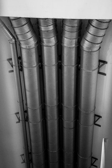 Four large industrial ducts run vertically up a wall, made of metal with ribbed textures. The ducts are evenly spaced and connected to a ceiling structure.