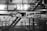 The image depicts industrial machinery in a factory setting. The machinery is complex, featuring various components and metal framework. The background includes a metal staircase, railings, and industrial lighting, giving the scene an industrial and mechanical atmosphere.