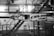 The image depicts industrial machinery in a factory setting. The machinery is complex, featuring various components and metal framework. The background includes a metal staircase, railings, and industrial lighting, giving the scene an industrial and mechanical atmosphere.
