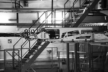 The image depicts industrial machinery in a factory setting. The machinery is complex, featuring various components and metal framework. The background includes a metal staircase, railings, and industrial lighting, giving the scene an industrial and mechanical atmosphere.