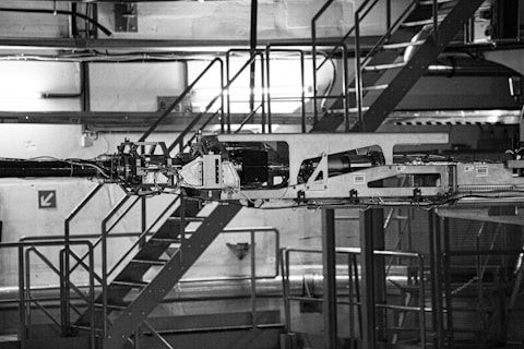 The image depicts industrial machinery in a factory setting. The machinery is complex, featuring various components and metal framework. The background includes a metal staircase, railings, and industrial lighting, giving the scene an industrial and mechanical atmosphere.