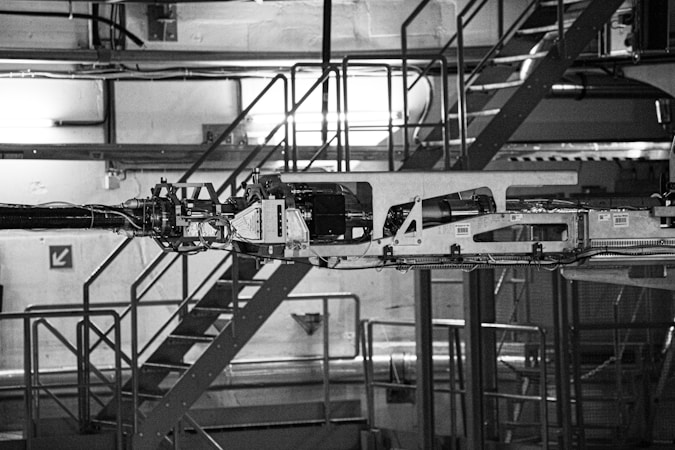 The image depicts industrial machinery in a factory setting. The machinery is complex, featuring various components and metal framework. The background includes a metal staircase, railings, and industrial lighting, giving the scene an industrial and mechanical atmosphere.