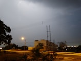 A lightning flash illuminates a cloudy night sky above a suburban landscape. The scene includes a street with some vegetation and a small building in the middle distance. Power lines run across the image, and a streetlight casts a yellow glow, creating a moody ambiance.