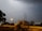 A lightning flash illuminates a cloudy night sky above a suburban landscape. The scene includes a street with some vegetation and a small building in the middle distance. Power lines run across the image, and a streetlight casts a yellow glow, creating a moody ambiance.