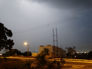 A lightning flash illuminates a cloudy night sky above a suburban landscape. The scene includes a street with some vegetation and a small building in the middle distance. Power lines run across the image, and a streetlight casts a yellow glow, creating a moody ambiance.
