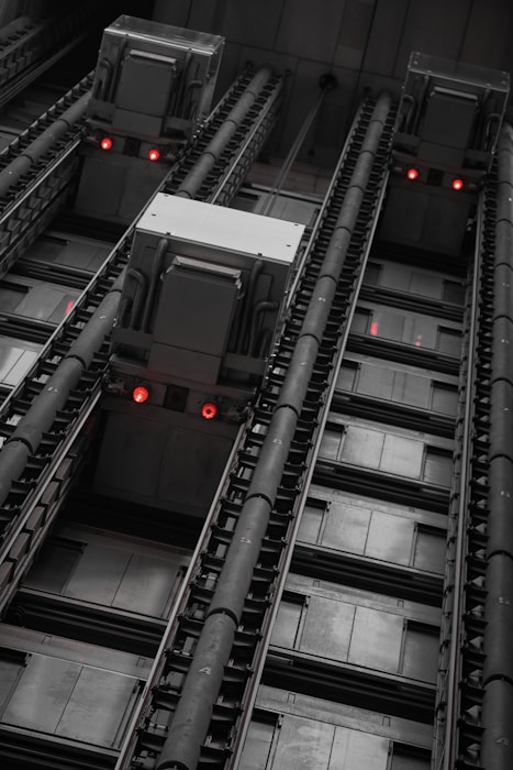 View of an industrial elevator system, featuring large mechanical components and rail tracks with red indicator lights. The perspective is from the bottom looking up, creating a sense of height and depth.