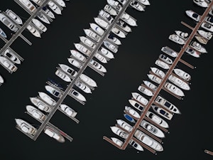 An aerial view of a marina with several rows of boats and yachts docked alongside wooden piers. The water appears dark, contrasting with the bright colors of the boats.