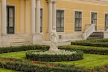 A detailed garden scene in front of a classical-style building with a yellow facade and black-framed windows. The garden is meticulously landscaped with trimmed hedges, patches of green grass, and red and white flowers. In the center is a marble statue of a human figure surrounded by lush greenery. The building features large columns and a decorative staircase leading to an entrance.