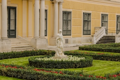 A detailed garden scene in front of a classical-style building with a yellow facade and black-framed windows. The garden is meticulously landscaped with trimmed hedges, patches of green grass, and red and white flowers. In the center is a marble statue of a human figure surrounded by lush greenery. The building features large columns and a decorative staircase leading to an entrance.