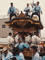 A group of people dressed in traditional attire are participating in a cultural festival. Several individuals are standing atop a decorated float, while others are playing musical instruments and wearing coordinated outfits with blue tones.