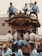 A group of people dressed in traditional attire are participating in a cultural festival. Several individuals are standing atop a decorated float, while others are playing musical instruments and wearing coordinated outfits with blue tones.
