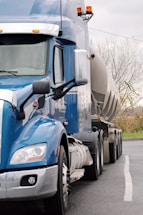 A blue semi-truck with a large tanker trailer is parked on a road. The truck features chrome accents and side mirrors, with the tanker extending towards the back. The background includes overcast skies and leafless trees, indicating a cold or late autumn setting.