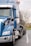 A blue semi-truck with a large tanker trailer is parked on a road. The truck features chrome accents and side mirrors, with the tanker extending towards the back. The background includes overcast skies and leafless trees, indicating a cold or late autumn setting.