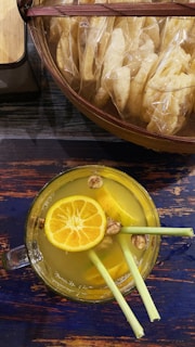 A glass cup filled with a bright yellow liquid garnished with a slice of orange, lemongrass stalks, and nuts. The drink is placed on a dark wooden table with hints of blue. Above the cup, a basket filled with packaged snacks is visible.