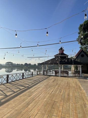 A wooden deck extends out towards a tranquil lake with a clear blue sky in the background. Strings of light bulbs are hung above the deck, connecting from one end to another. There is a gazebo-like structure with a black roof and white columns, under which several wooden tables and chairs are arranged. The railing along the edge of the deck is light-colored, creating a safe boundary between the deck and the water.
