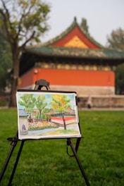 An easel stands on green grass, holding a watercolor painting of a traditional East Asian building with a red wall and green roof. In the background, the actual building is partially visible, surrounded by trees.
