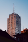 A tall building featuring Art Deco architectural style with an antenna tower on the top. The facade has intricate details and numerous windows reflecting sunlight, creating a warm glow. In the foreground, there are silhouettes of lower structures, suggesting an urban setting.