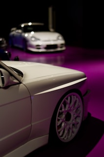 Two sleek white cars are showcased, positioned on a glossy purple floor. The foreground features a close-up of one car's side, highlighting its intricate wheel design. In the background, a second car is seen, adding depth to the scene with a dark ambient setting enhancing the vehicles.