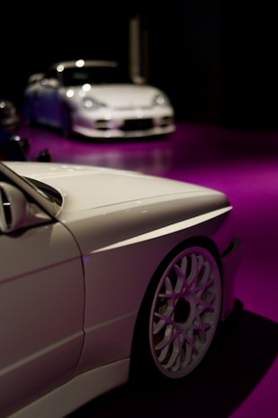 Two sleek white cars are showcased, positioned on a glossy purple floor. The foreground features a close-up of one car's side, highlighting its intricate wheel design. In the background, a second car is seen, adding depth to the scene with a dark ambient setting enhancing the vehicles.