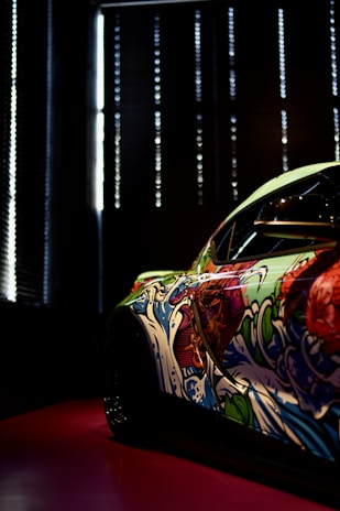 A car with an artistic and colorful graffiti design on its side, featuring vibrant waves and abstract patterns. The scene is dimly lit, with light coming through vertical blinds in the background, creating a dramatic effect.