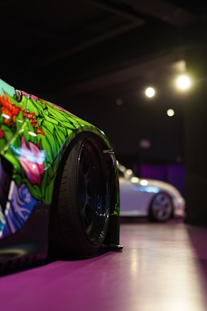 A close-up view of a car with intricate and colorful floral designs on the body, focusing on the wheel. In the background, another car, mostly white, is visible under low lighting, creating a dim and atmospheric setting.