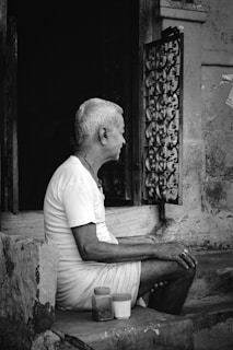 An elderly person with short white hair and a white shirt sits on a stone step in front of a weathered doorway. Their legs are bent, and they appear to be looking out towards an ornate iron grille fixed to the wall. Two small containers rest near their feet, providing a contrast in size and shape against the rugged stone.