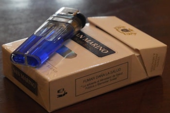 A blue translucent lighter rests on top of a closed pack of San Marino cigarettes. The cigarette pack has a health warning label in Spanish and is placed on a dark surface.