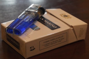 A blue translucent lighter rests on top of a closed pack of San Marino cigarettes. The cigarette pack has a health warning label in Spanish and is placed on a dark surface.