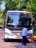 A man wearing a blue shirt is standing next to a parked white bus with visible wear near the front bumper. The bus is stopped on the side of a tree-lined street, and the man appears to be cleaning or inspecting the front. Sunlight filters through the lush green trees, casting shadows on the road.