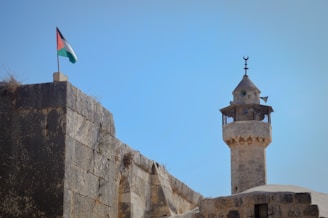 A historic stone structure with a tall minaret rises against a clear blue sky. A flag is prominently displayed on top of a sturdy stone wall adjacent to the minaret.