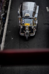 A top-down view of a three-wheeled taxi, commonly known as a tuk-tuk, driving on a dark asphalt road. The vehicle has a metallic roof, a prominent 'TAXI' sign on top, and a yellow license plate. The road has a white line on the left side and is surrounded by concrete barriers.