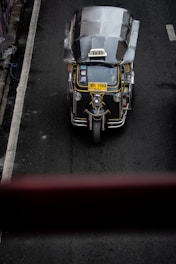 A top-down view of a three-wheeled taxi, commonly known as a tuk-tuk, driving on a dark asphalt road. The vehicle has a metallic roof, a prominent 'TAXI' sign on top, and a yellow license plate. The road has a white line on the left side and is surrounded by concrete barriers.