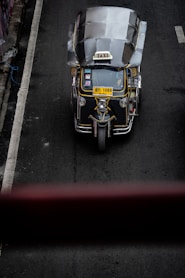 A top-down view of a three-wheeled taxi, commonly known as a tuk-tuk, driving on a dark asphalt road. The vehicle has a metallic roof, a prominent 'TAXI' sign on top, and a yellow license plate. The road has a white line on the left side and is surrounded by concrete barriers.