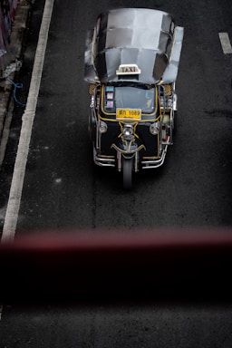 A top-down view of a three-wheeled taxi, commonly known as a tuk-tuk, driving on a dark asphalt road. The vehicle has a metallic roof, a prominent 'TAXI' sign on top, and a yellow license plate. The road has a white line on the left side and is surrounded by concrete barriers.