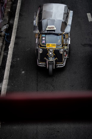 A top-down view of a three-wheeled taxi, commonly known as a tuk-tuk, driving on a dark asphalt road. The vehicle has a metallic roof, a prominent 'TAXI' sign on top, and a yellow license plate. The road has a white line on the left side and is surrounded by concrete barriers.