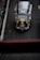 A top-down view of a three-wheeled taxi, commonly known as a tuk-tuk, driving on a dark asphalt road. The vehicle has a metallic roof, a prominent 'TAXI' sign on top, and a yellow license plate. The road has a white line on the left side and is surrounded by concrete barriers.
