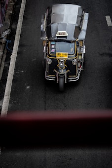 A top-down view of a three-wheeled taxi, commonly known as a tuk-tuk, driving on a dark asphalt road. The vehicle has a metallic roof, a prominent 'TAXI' sign on top, and a yellow license plate. The road has a white line on the left side and is surrounded by concrete barriers.