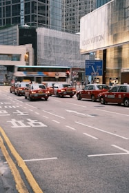 A busy urban street scene with several red taxis lined up at a traffic signal in front of a luxury store with glass buildings in the background. The atmosphere is bustling with movement as a bus passes by quickly, creating a blur effect. The street markings include both English and Chinese characters.