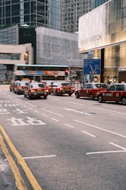 A busy urban street scene with several red taxis lined up at a traffic signal in front of a luxury store with glass buildings in the background. The atmosphere is bustling with movement as a bus passes by quickly, creating a blur effect. The street markings include both English and Chinese characters.