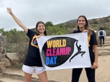 Two people are holding a sign that reads 'World Cleanup Day' in a natural outdoor setting. They are smiling and wearing event-themed t-shirts. There is a stone wall and greenery in the background, and additional people can be seen walking up steps.