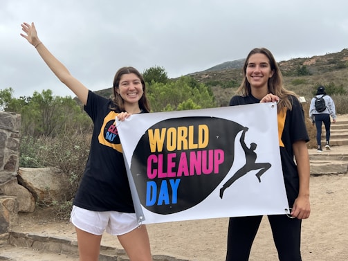 Two people are holding a sign that reads 'World Cleanup Day' in a natural outdoor setting. They are smiling and wearing event-themed t-shirts. There is a stone wall and greenery in the background, and additional people can be seen walking up steps.